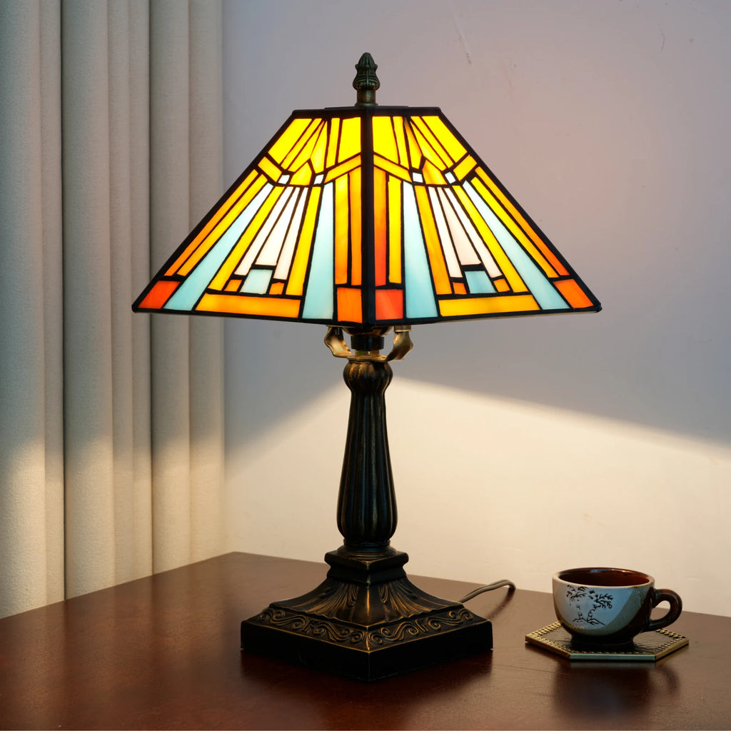 Stained glass pyramid lamp lit on a dark wood table, amber and sky-blue panels, warm glow on the wall